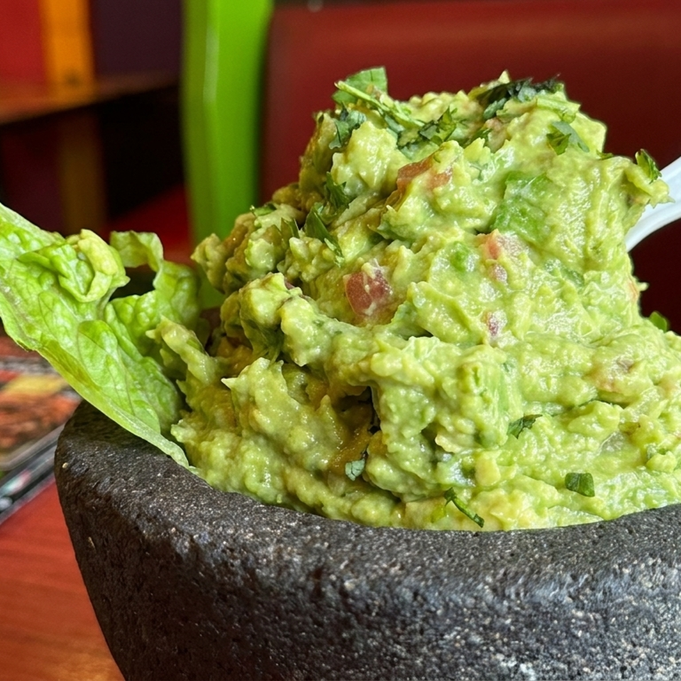 Fresh guacamole garnished with cilantro and tomatoes in a black stone bowl, served with lettuce and a spoon at a colorful restaurant table.